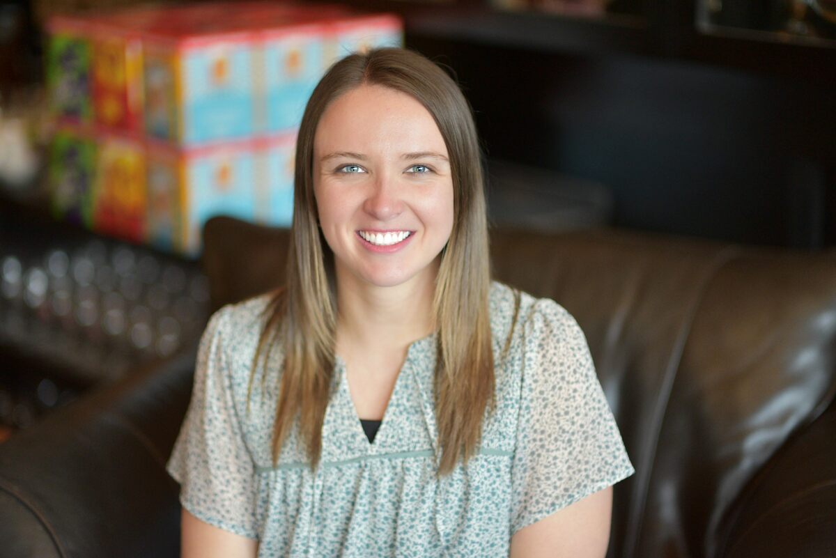 Cara Metts, in light blue blouse, sitting on brown couch.
